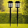 Two black solar garden lights with white paneled tops stand in a vibrant green lawn with hedges and trees.